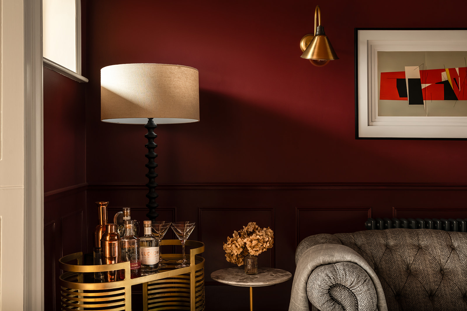 Moody red snug with brass sconce, sculptural black table lamp and drinks trolley, demonstrating ambient lighting in a period home by Studio 3 Creatives.