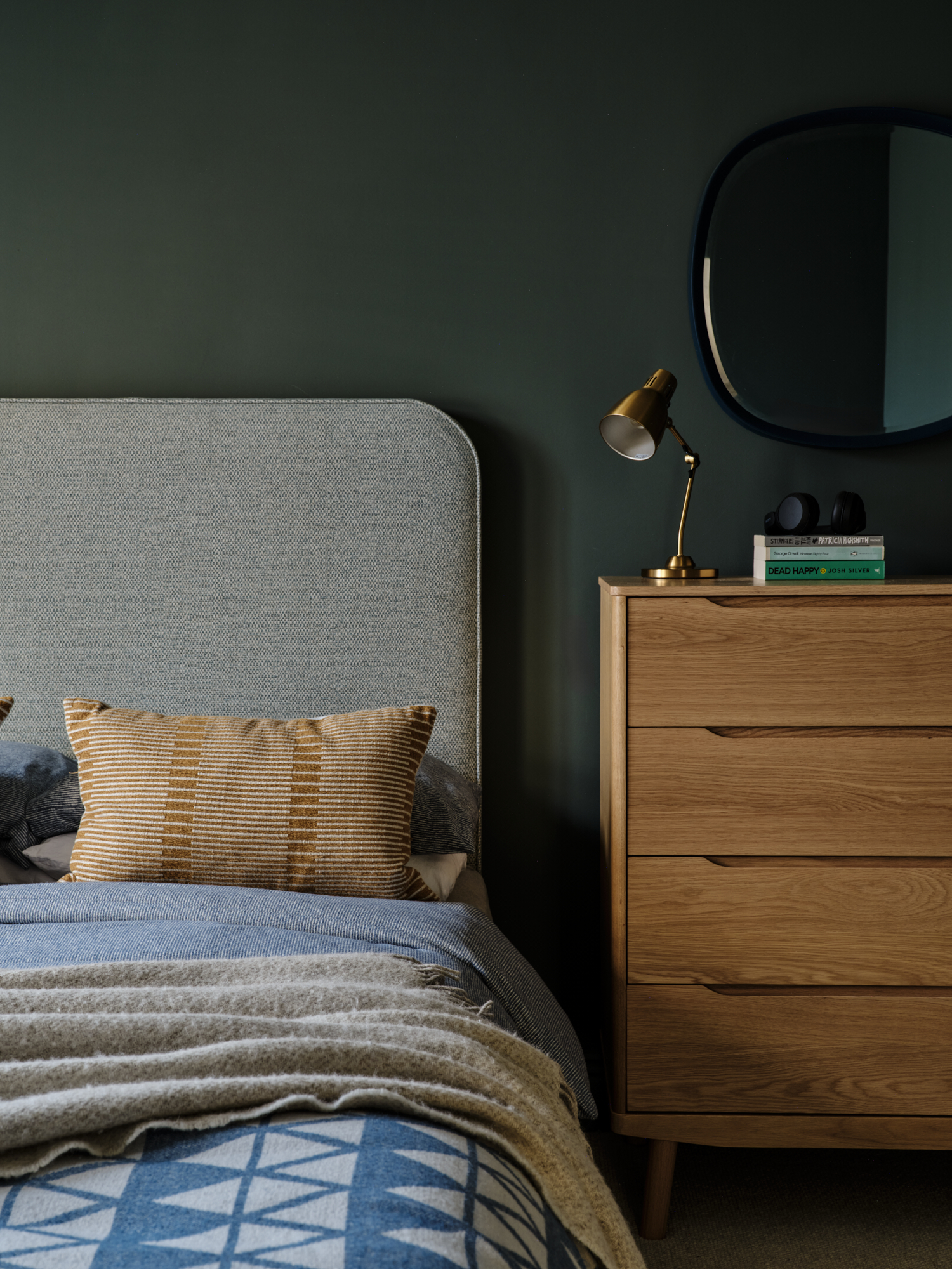 Forest green teenage bedroom with upholstered headboard, oak chest of drawers with brass lamp and round navy mirror, interior design London